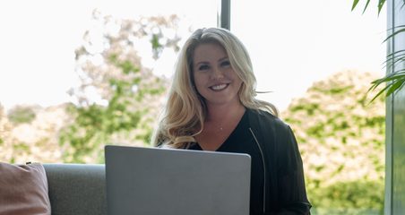 woman smiling in front of the camera while sitting on a couch and working with a laptop