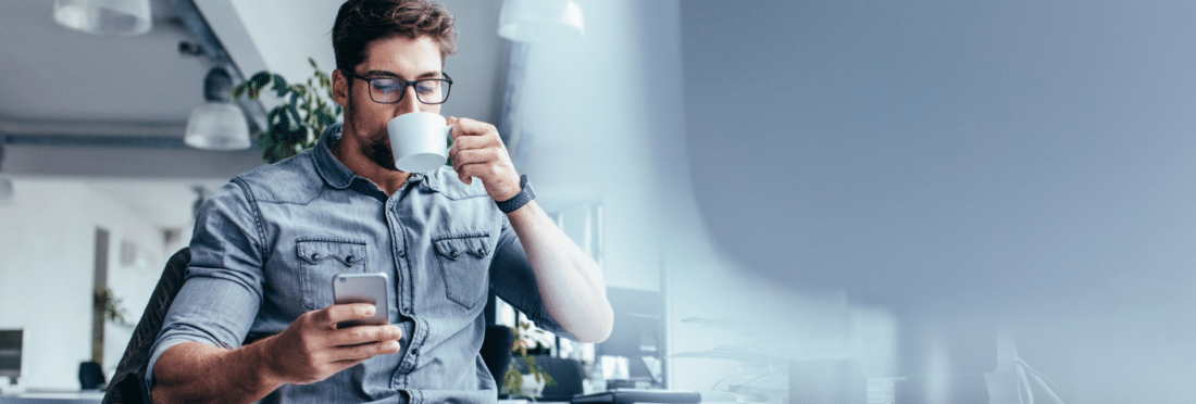 Man looking at iPhone while drinking coffee at his desk